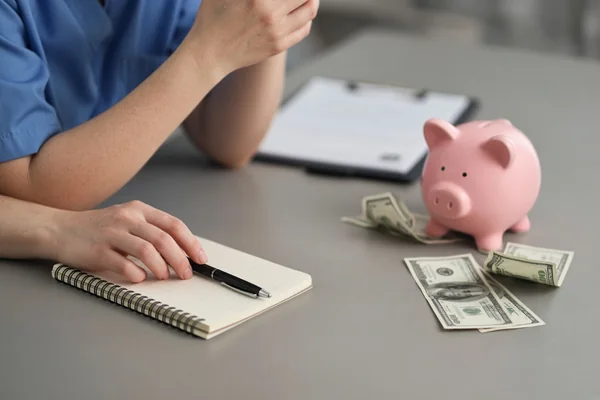 A nurse in blue scrubs at a table with a notepad and pen, a clipboard behind, and a piggy bank beside folded banknotes.