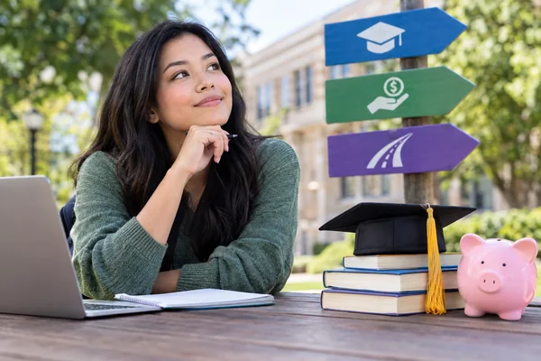 A woman at an outdoor table with a laptop and notebook, looking toward three directional signs marked with a graduation cap, a hand holding a coin, and a road.