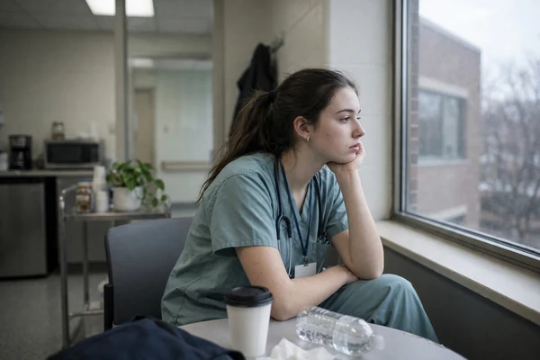 A nurse in scrubs with a stethoscope sitting in a staff break room beside a window, chin resting on one hand, looking out.